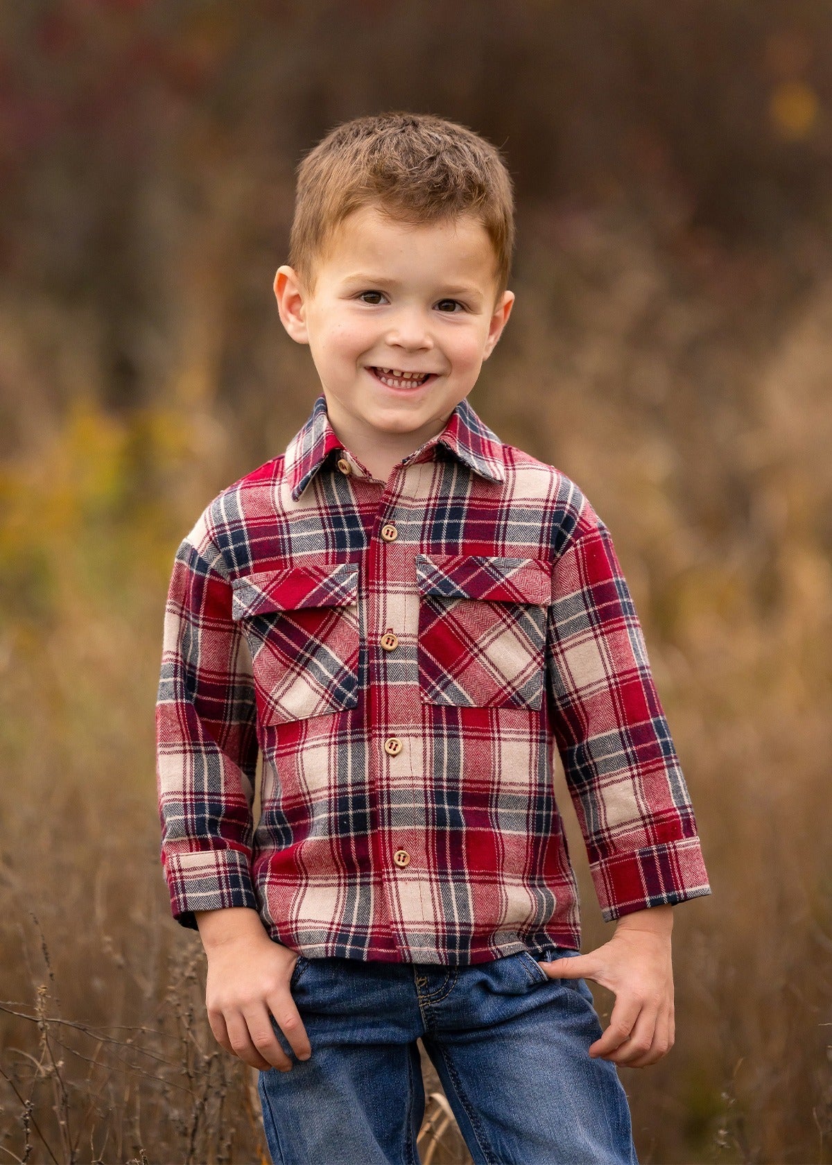 A young boy with short brown hair smiles outdoors, wearing the Beckett and Bear Henry Jacket. The background is softly blurred with brown and green tones, highlighting a natural setting.