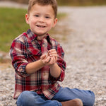 A young boy wearing the Beckett and Bear Henry Jacket sits cross-legged on a gravel path, smiling at the camera with small rocks in his hands, grass and blurred trees in the background.