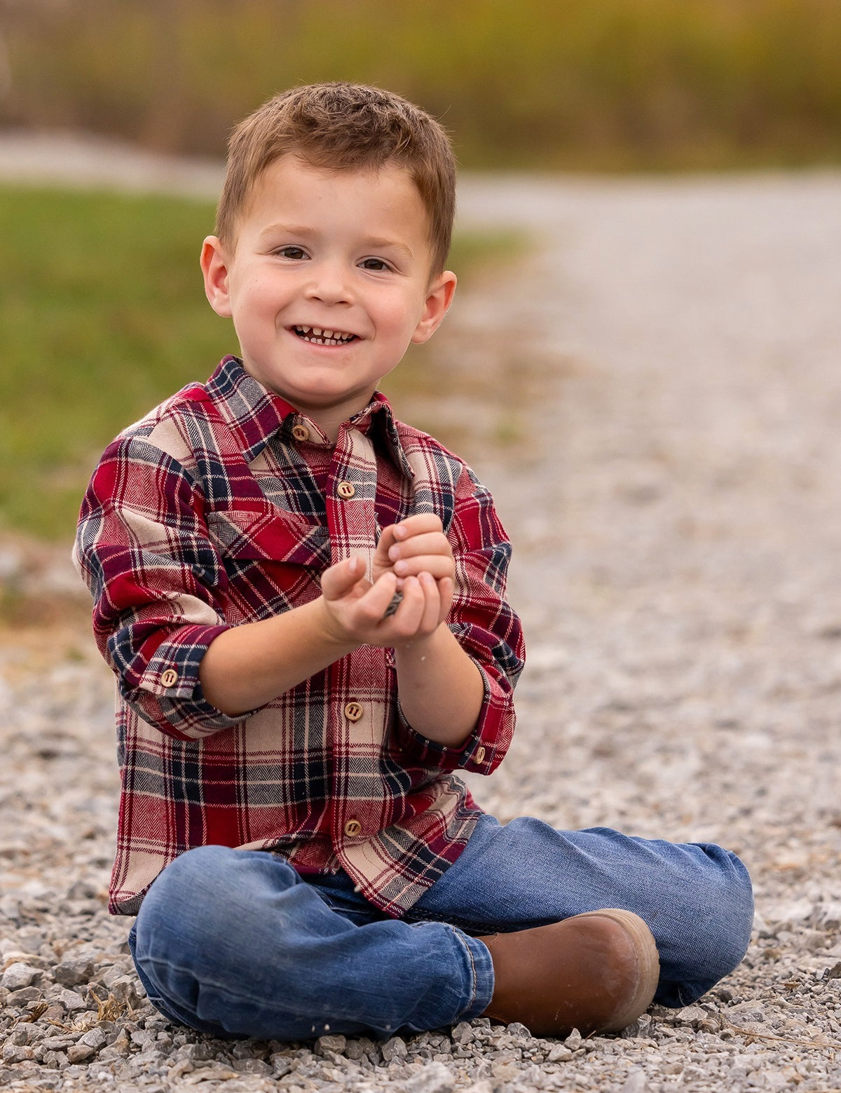 A young boy wearing the Beckett and Bear Henry Jacket sits cross-legged on a gravel path, smiling at the camera with small rocks in his hands, grass and blurred trees in the background.
