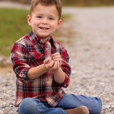 A young boy wearing the Beckett and Bear Henry Jacket sits cross-legged on a gravel path, smiling at the camera with small rocks in his hands, grass and blurred trees in the background.