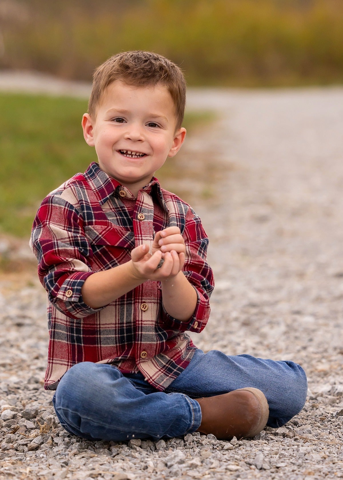 A young boy wearing the Beckett and Bear Henry Jacket sits cross-legged on a gravel path, smiling at the camera with small rocks in his hands, grass and blurred trees in the background.