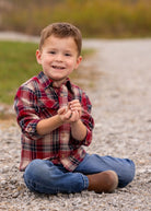 A young boy wearing the Beckett and Bear Henry Jacket sits cross-legged on a gravel path, smiling at the camera with small rocks in his hands, grass and blurred trees in the background.
