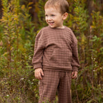 A young child with short light brown hair wears the Theo Two Piece Set from Beckett and Bear, smiling while standing in a grassy outdoor area surrounded by tall green plants.