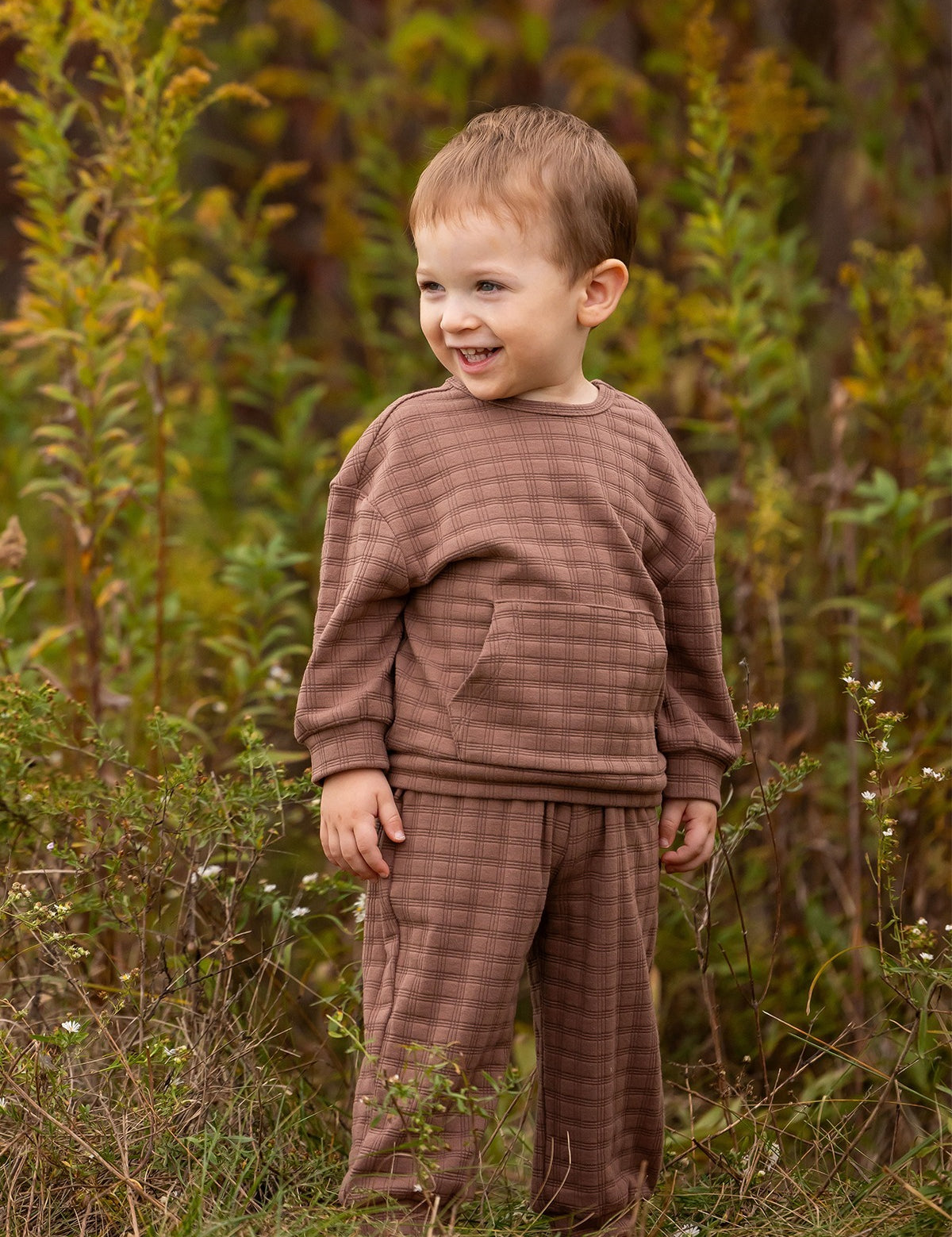 A young child with short light brown hair wears the Theo Two Piece Set from Beckett and Bear, smiling while standing in a grassy outdoor area surrounded by tall green plants.