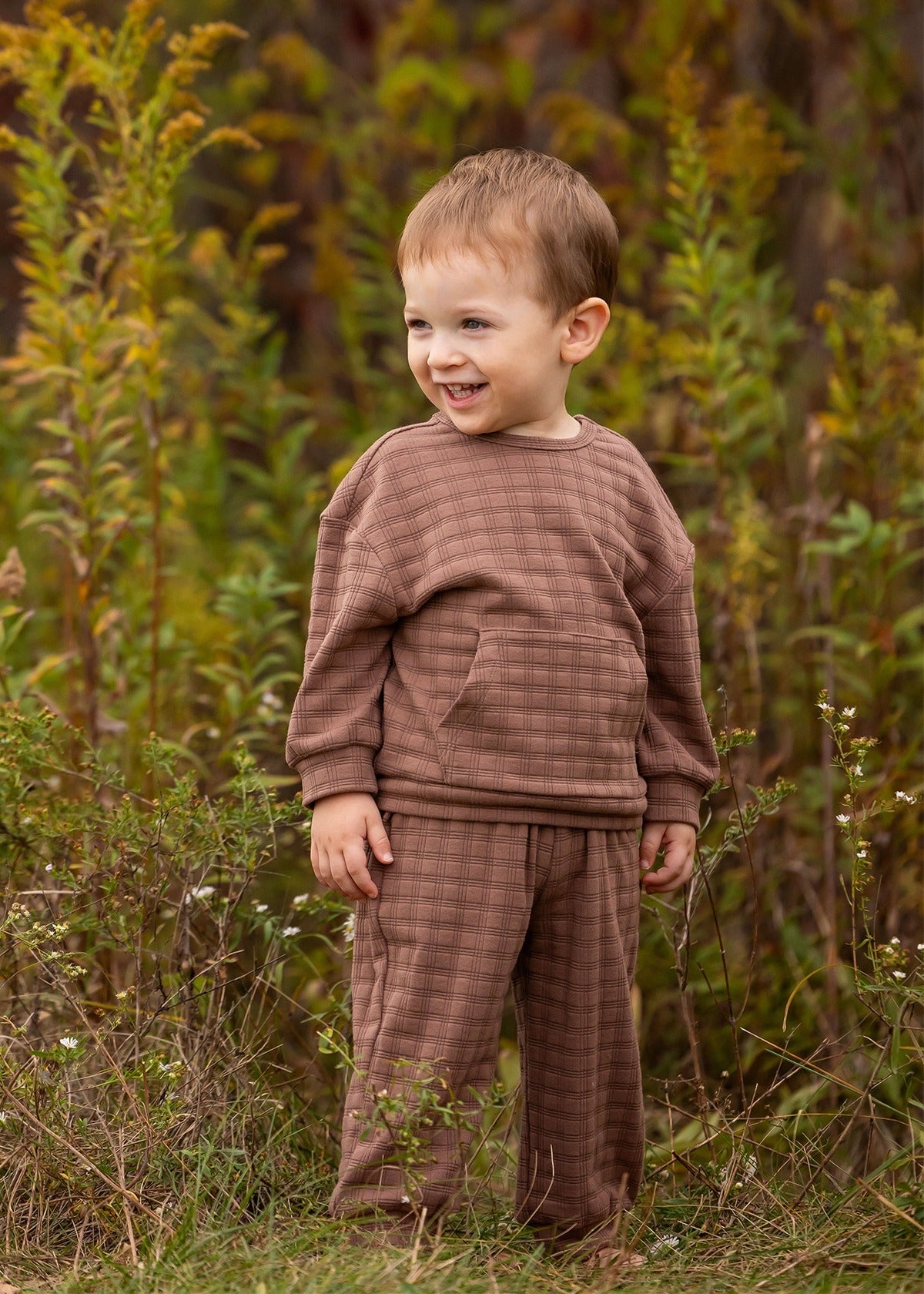A young child with short light brown hair wears the Theo Two Piece Set from Beckett and Bear, smiling while standing in a grassy outdoor area surrounded by tall green plants.