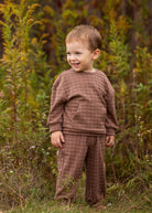 A young child with short light brown hair wears the Theo Two Piece Set from Beckett and Bear, smiling while standing in a grassy outdoor area surrounded by tall green plants.