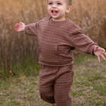 A young child wearing the Beckett and Bear Theo Two Piece Set and white sneakers walks with arms outstretched, smiling outdoors on grass with tall, dry grass in the background.