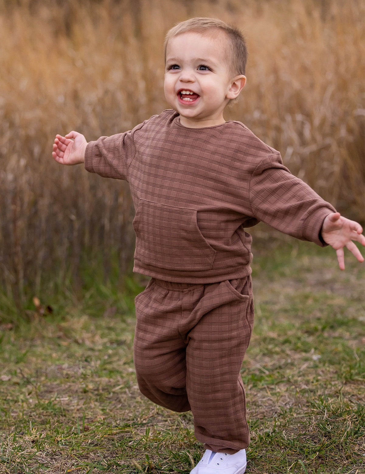 A young child wearing the Beckett and Bear Theo Two Piece Set and white sneakers walks with arms outstretched, smiling outdoors on grass with tall, dry grass in the background.