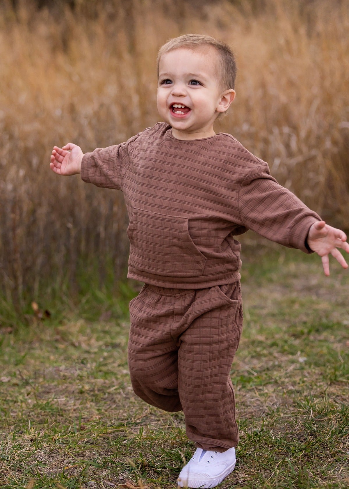 A young child wearing the Beckett and Bear Theo Two Piece Set and white sneakers walks with arms outstretched, smiling outdoors on grass with tall, dry grass in the background.
