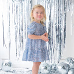 A young girl wears the Isobella and Chloe Starry Blue Glitter Dress and beige shoes, smiling amid silver tinsel streamers and disco balls on a white floor for a festive party look.