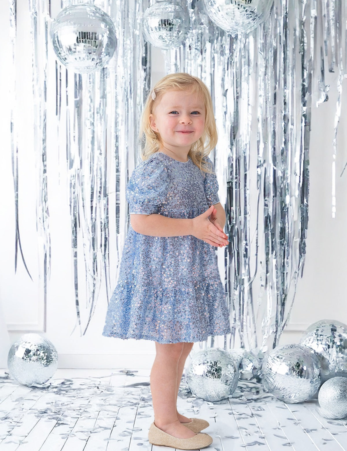 A young girl wears the Isobella and Chloe Starry Blue Glitter Dress and beige shoes, smiling amid silver tinsel streamers and disco balls on a white floor for a festive party look.