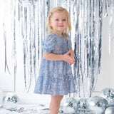 A young girl wears the Isobella and Chloe Starry Blue Glitter Dress and beige shoes, smiling amid silver tinsel streamers and disco balls on a white floor for a festive party look.