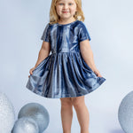 A young blonde girl smiles in the Isobella and Chloe Starry Blue Velvet Dress, holding the sides of her special occasion outfit, surrounded by large glittery silver spheres on a light blue background.