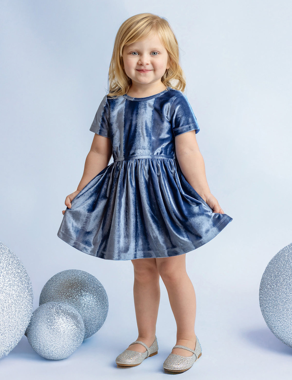 A young blonde girl smiles in the Isobella and Chloe Starry Blue Velvet Dress, holding the sides of her special occasion outfit, surrounded by large glittery silver spheres on a light blue background.