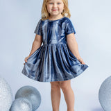 A young blonde girl smiles in the Isobella and Chloe Starry Blue Velvet Dress, holding the sides of her special occasion outfit, surrounded by large glittery silver spheres on a light blue background.