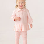A smiling toddler with light hair in pigtails wears the Serena Two Piece Set by Mabel and Honey—a pale pink girls' outfit with a collared top and matching leggings—paired with white sneakers, standing indoors against a plain background.
