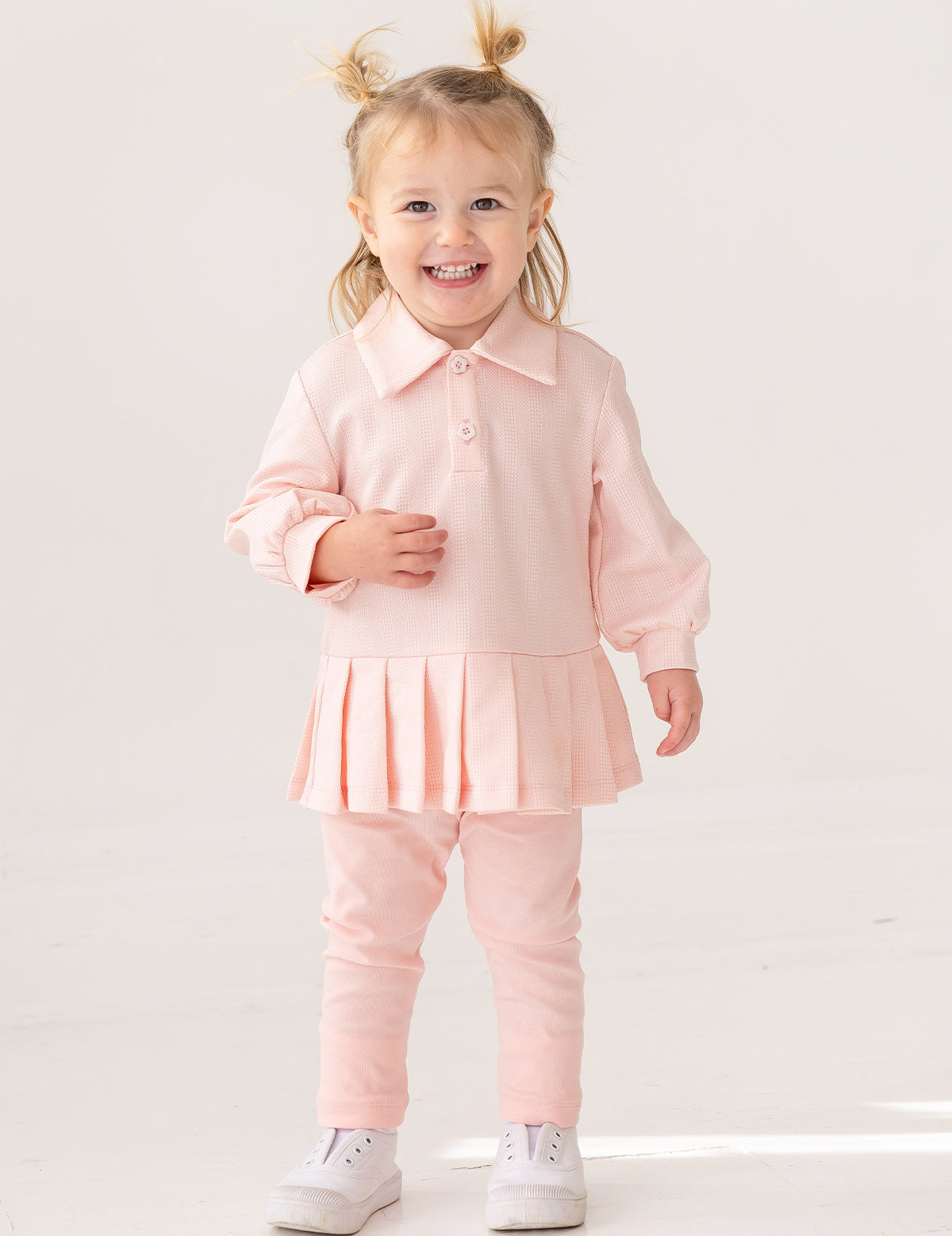 A smiling toddler with light hair in pigtails wears the Serena Two Piece Set by Mabel and Honey—a pale pink girls' outfit with a collared top and matching leggings—paired with white sneakers, standing indoors against a plain background.