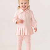 A smiling toddler with light hair in pigtails wears the Serena Two Piece Set by Mabel and Honey—a pale pink girls' outfit with a collared top and matching leggings—paired with white sneakers, standing indoors against a plain background.