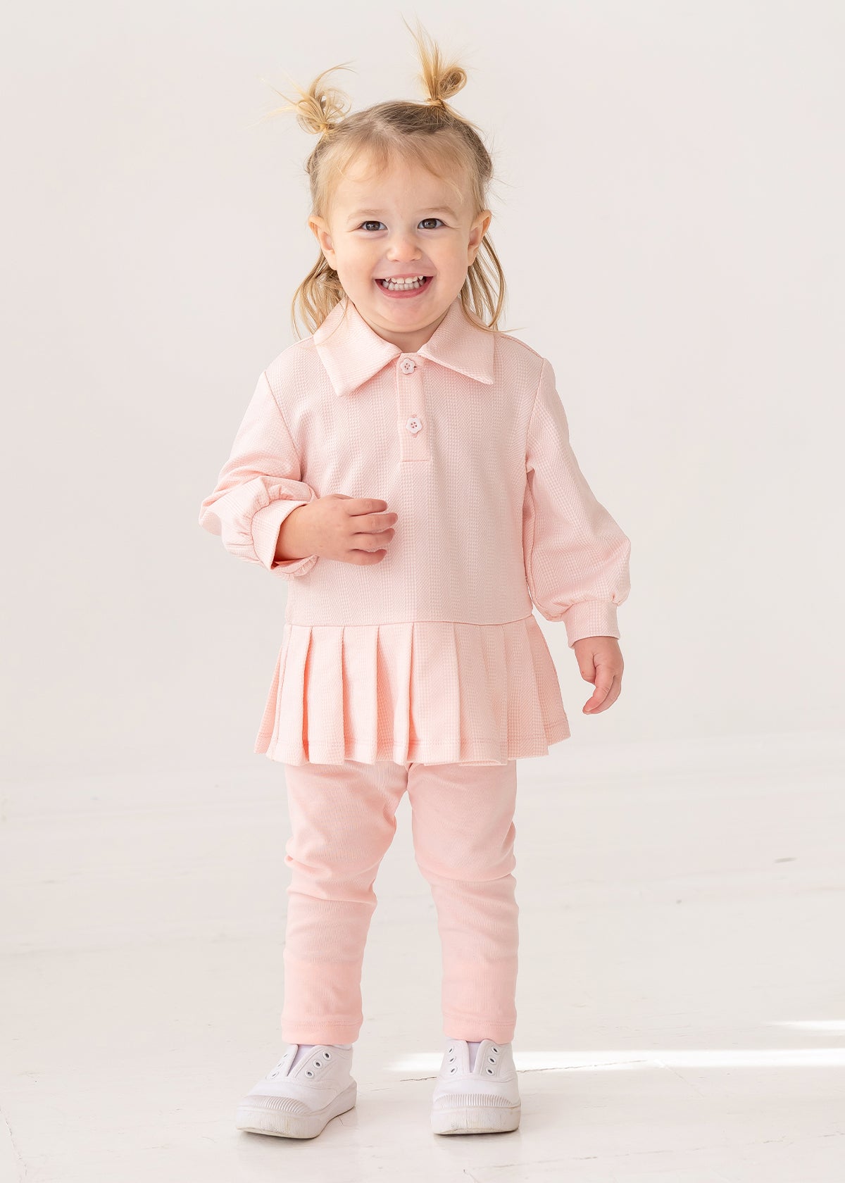 A smiling toddler with light hair in pigtails wears the Serena Two Piece Set by Mabel and Honey—a pale pink girls' outfit with a collared top and matching leggings—paired with white sneakers, standing indoors against a plain background.