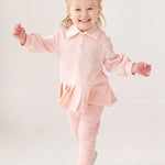 A young child with light hair in two buns smiles energetically, wearing the Serena Two Piece Set by Mabel and Honey—a light pink long-sleeve top with pleated hem skirt, matching pants, and white shoes against a plain white background.
