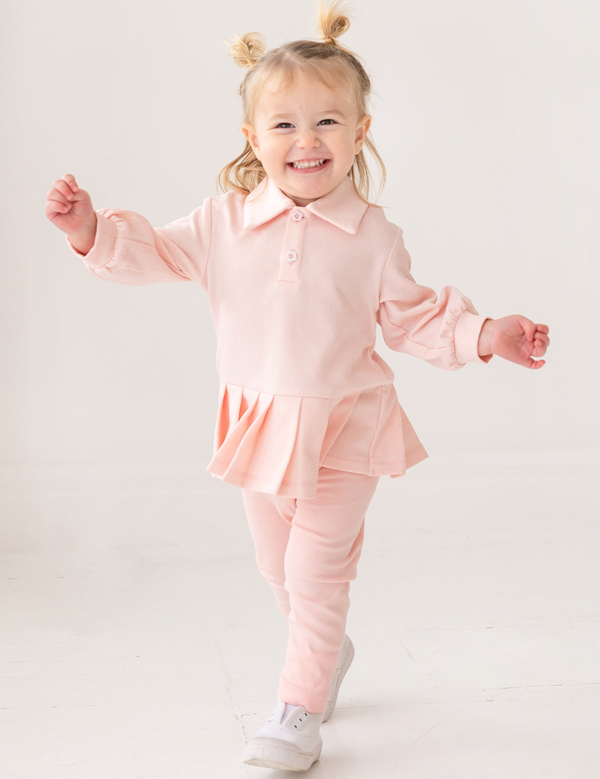 A young child with light hair in two buns smiles energetically, wearing the Serena Two Piece Set by Mabel and Honey—a light pink long-sleeve top with pleated hem skirt, matching pants, and white shoes against a plain white background.