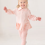 A young child with light hair in two buns smiles energetically, wearing the Serena Two Piece Set by Mabel and Honey—a light pink long-sleeve top with pleated hem skirt, matching pants, and white shoes against a plain white background.