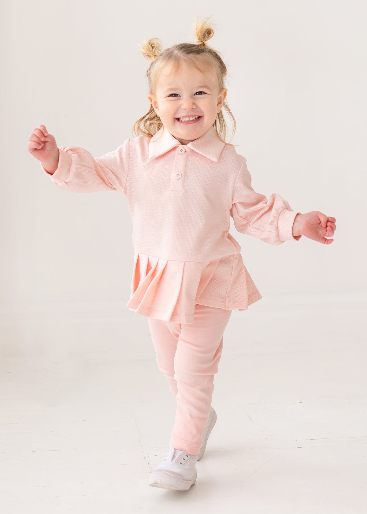 A young child with light hair in two buns smiles energetically, wearing the Serena Two Piece Set by Mabel and Honey—a light pink long-sleeve top with pleated hem skirt, matching pants, and white shoes against a plain white background.