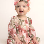 A baby smiles in Mabel and Honey’s Bow-tiful Romper—an adorable long-sleeve romper with a pink and green bow print, paired with a matching headband—set against a light, plain background.