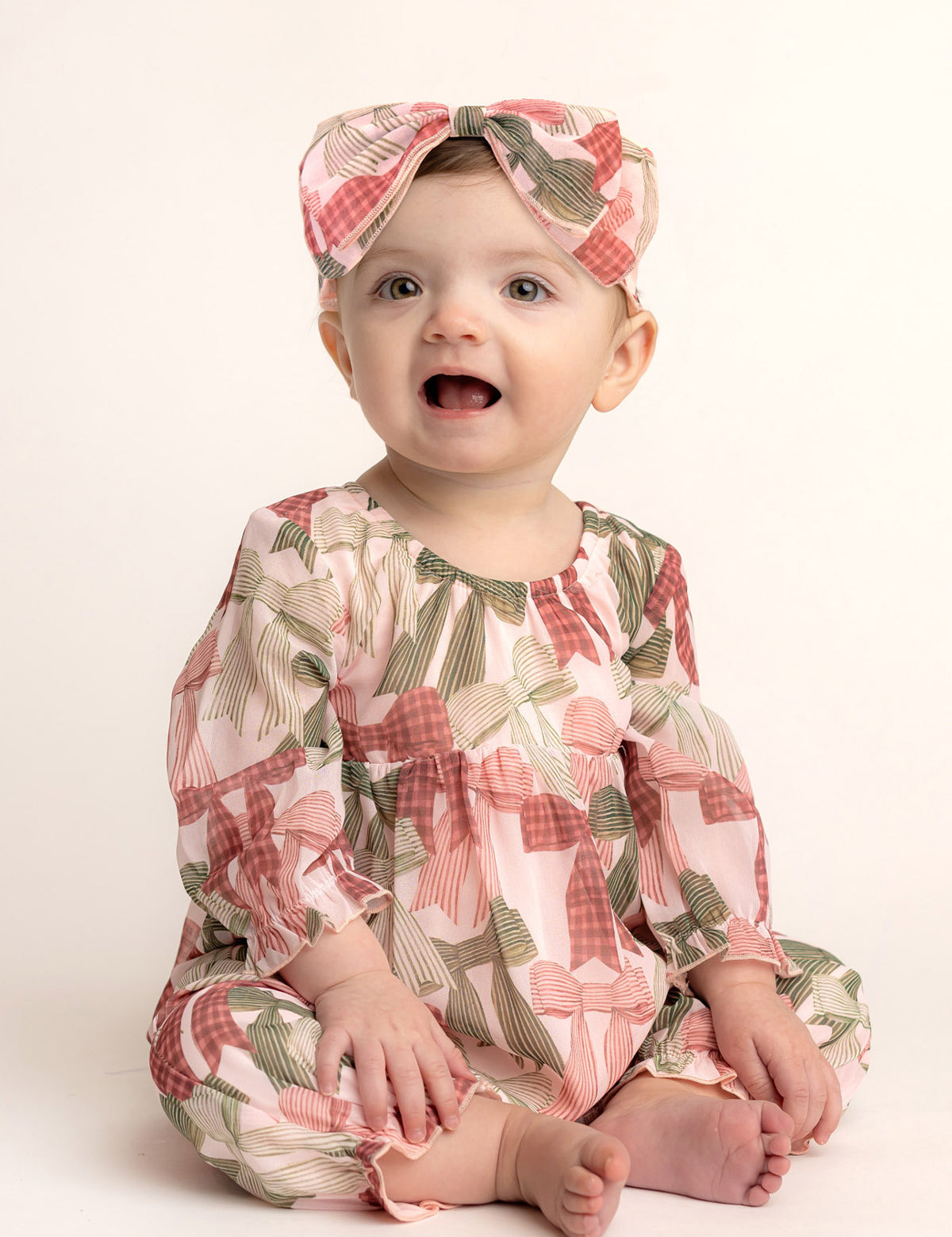 A baby smiles in Mabel and Honey’s Bow-tiful Romper—an adorable long-sleeve romper with a pink and green bow print, paired with a matching headband—set against a light, plain background.