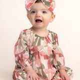 A baby smiles in Mabel and Honey’s Bow-tiful Romper—an adorable long-sleeve romper with a pink and green bow print, paired with a matching headband—set against a light, plain background.