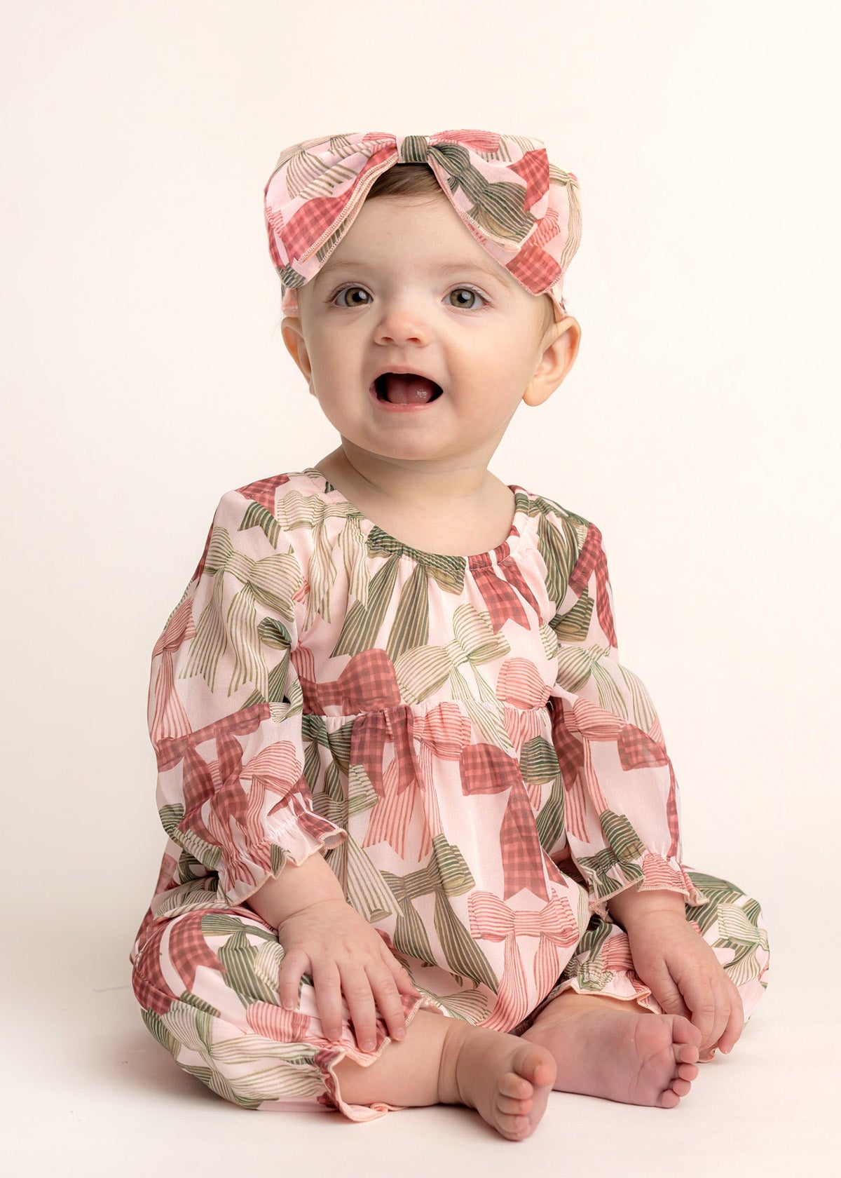 A baby smiles in Mabel and Honey’s Bow-tiful Romper—an adorable long-sleeve romper with a pink and green bow print, paired with a matching headband—set against a light, plain background.