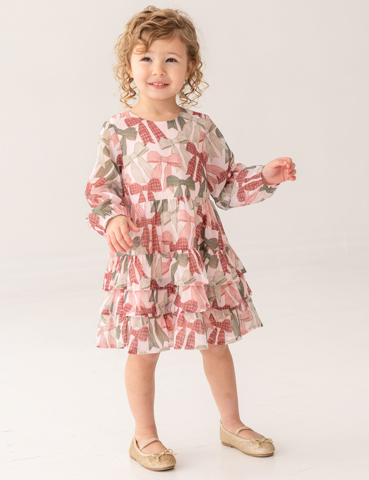 A young child with curly hair wears the Mabel and Honey Bow-tiful Dress, featuring a pink and green geometric pattern, paired with gold shoes, standing and smiling against a plain light background.