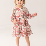 A young child with curly hair wears the Mabel and Honey Bow-tiful Dress, featuring a pink and green geometric pattern, paired with gold shoes, standing and smiling against a plain light background.