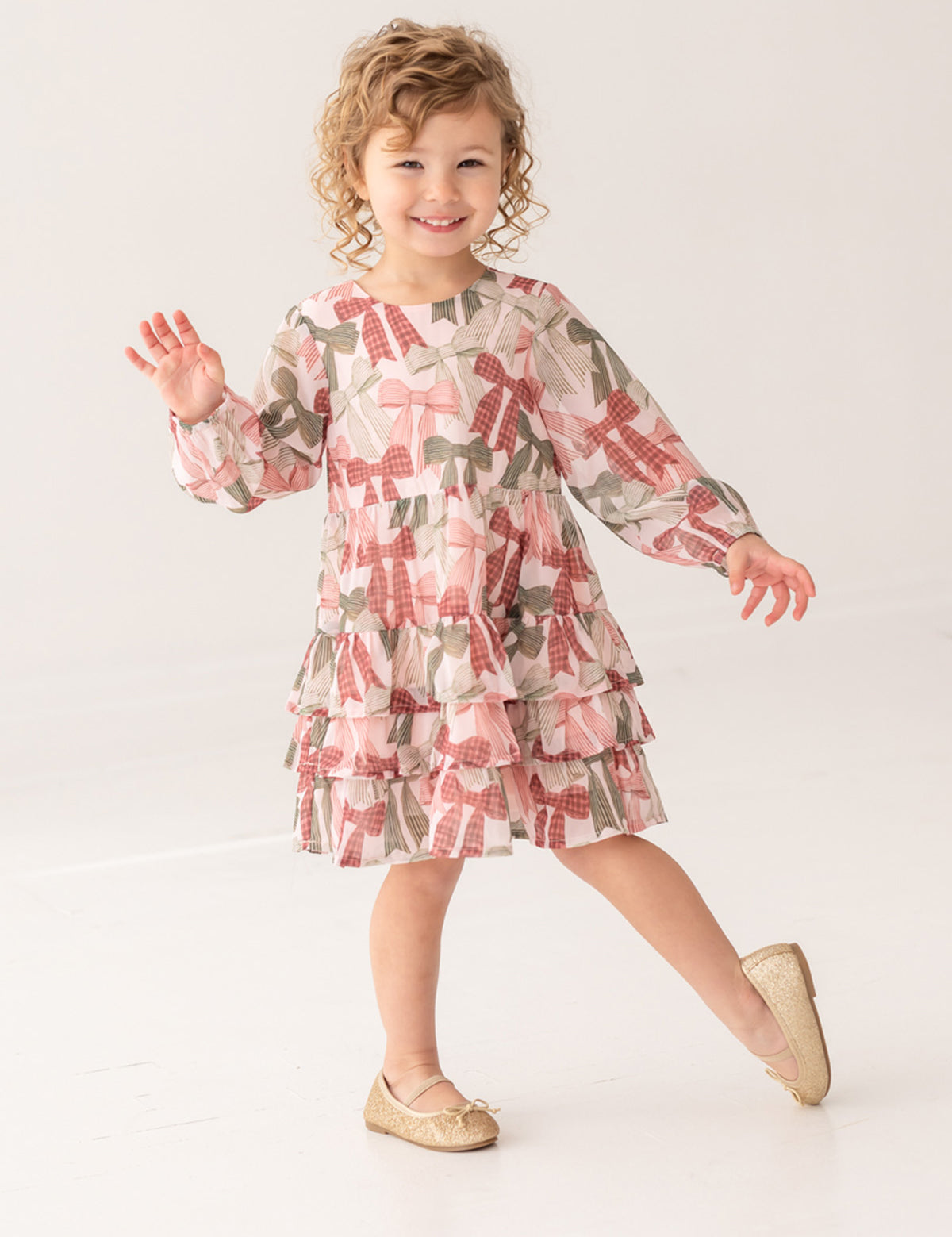 A curly-haired girl, smiling and waving, poses on one foot in a bright studio wearing the Bow-tiful Dress by Mabel and Honey with pink and olive tiered ruffles, paired with gold flats for a charming look.