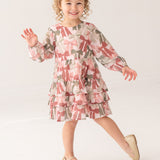 A curly-haired girl, smiling and waving, poses on one foot in a bright studio wearing the Bow-tiful Dress by Mabel and Honey with pink and olive tiered ruffles, paired with gold flats for a charming look.