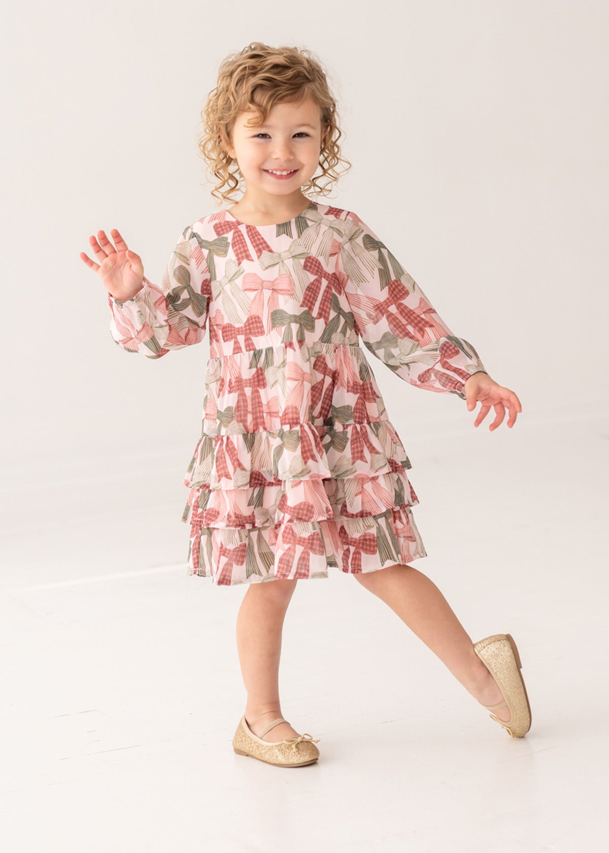 A curly-haired girl, smiling and waving, poses on one foot in a bright studio wearing the Bow-tiful Dress by Mabel and Honey with pink and olive tiered ruffles, paired with gold flats for a charming look.