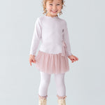 A young girl with curly hair and a bow headband is smiling, wearing the Isobella and Chloe Ballerina Two Piece Pant Set with a tulle skirt and matching leggings, paired with beige boots, standing on a white platform against a plain background.