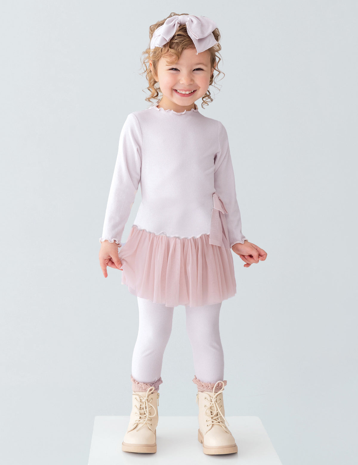 A young girl with curly hair and a bow headband is smiling, wearing the Isobella and Chloe Ballerina Two Piece Pant Set with a tulle skirt and matching leggings, paired with beige boots, standing on a white platform against a plain background.