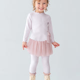A young girl with curly hair and a bow headband is smiling, wearing the Isobella and Chloe Ballerina Two Piece Pant Set with a tulle skirt and matching leggings, paired with beige boots, standing on a white platform against a plain background.