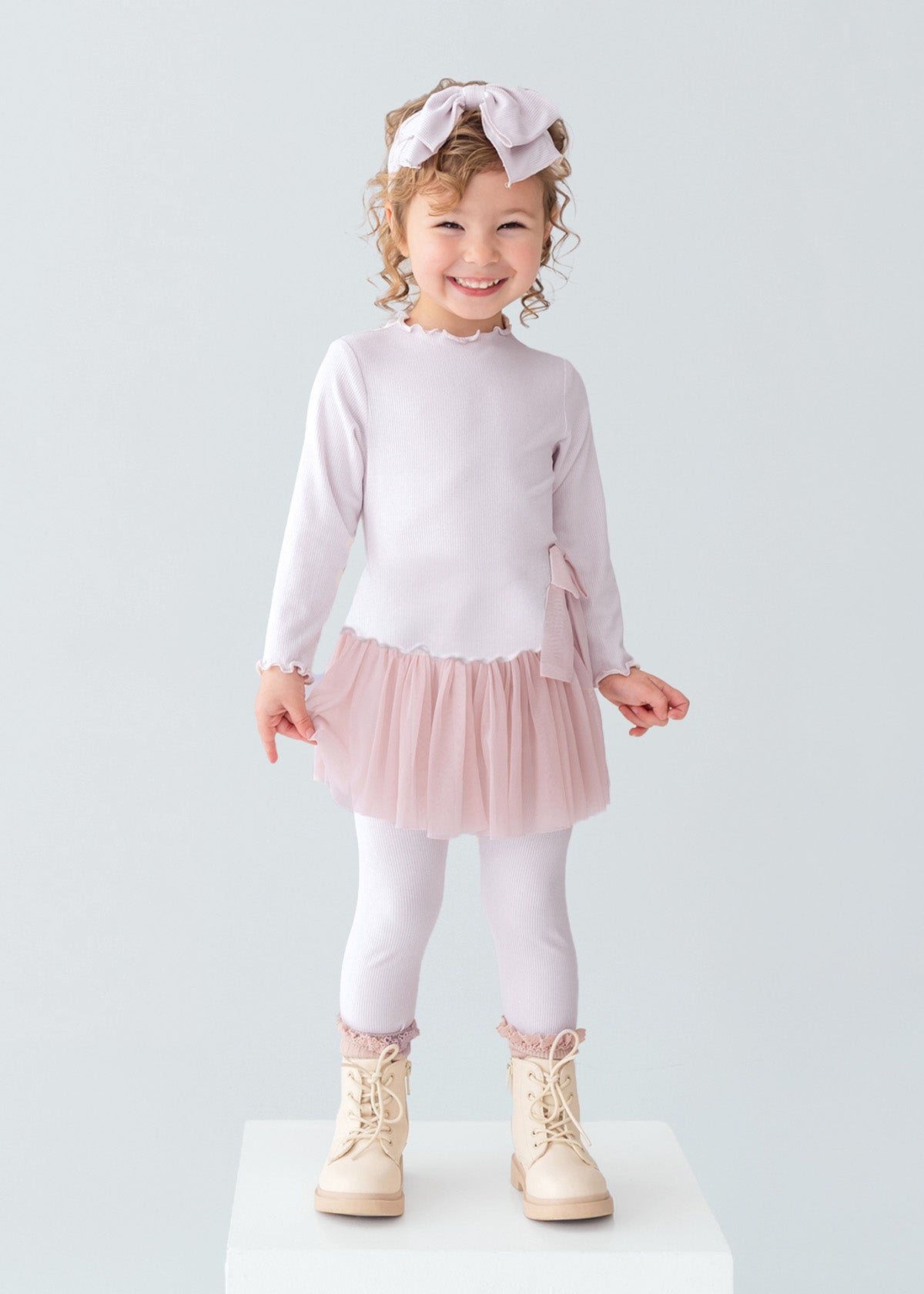A young girl with curly hair and a bow headband is smiling, wearing the Isobella and Chloe Ballerina Two Piece Pant Set with a tulle skirt and matching leggings, paired with beige boots, standing on a white platform against a plain background.