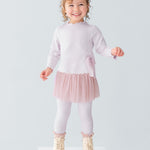 A young girl smiles on a white platform, wearing the Isobella and Chloe Ballerina Two Piece Pant Set with a tulle skirt, ribbed leggings, cream boots, and a large bow headband. The background is plain light gray.