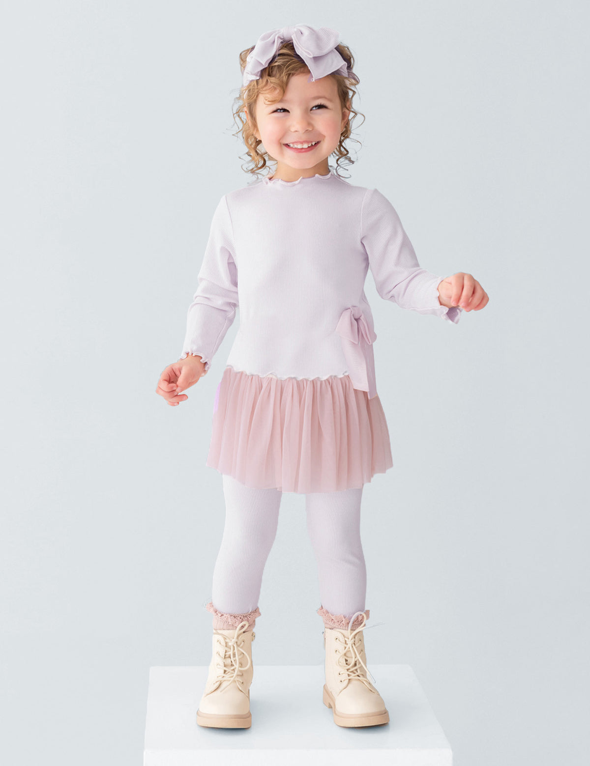 A young girl smiles on a white platform, wearing the Isobella and Chloe Ballerina Two Piece Pant Set with a tulle skirt, ribbed leggings, cream boots, and a large bow headband. The background is plain light gray.