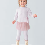 A young girl smiles on a white platform, wearing the Isobella and Chloe Ballerina Two Piece Pant Set with a tulle skirt, ribbed leggings, cream boots, and a large bow headband. The background is plain light gray.