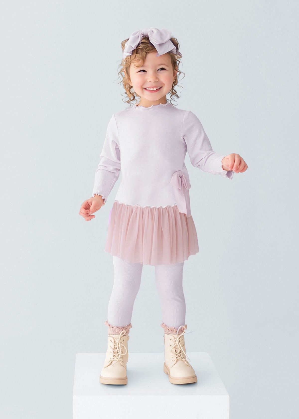 A young girl smiles on a white platform, wearing the Isobella and Chloe Ballerina Two Piece Pant Set with a tulle skirt, ribbed leggings, cream boots, and a large bow headband. The background is plain light gray.