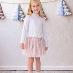 A young girl beams indoors in the Isobella and Chloe Ballerina Two Piece Set—featuring a white long-sleeve top and light pink tulle skirt—paired with white boots and lace socks, with paper tree decorations hanging behind her.