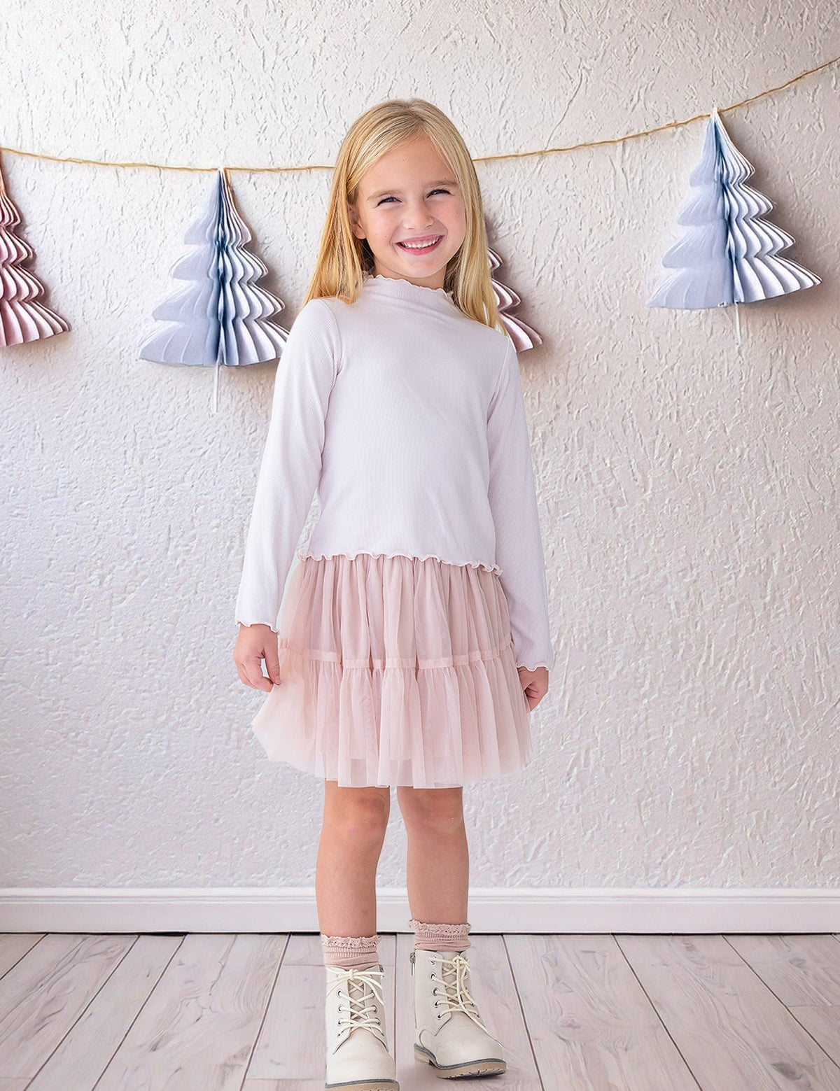 A young girl beams indoors in the Isobella and Chloe Ballerina Two Piece Set—featuring a white long-sleeve top and light pink tulle skirt—paired with white boots and lace socks, with paper tree decorations hanging behind her.