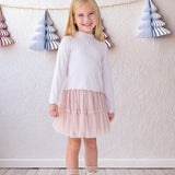 A young girl beams indoors in the Isobella and Chloe Ballerina Two Piece Set—featuring a white long-sleeve top and light pink tulle skirt—paired with white boots and lace socks, with paper tree decorations hanging behind her.