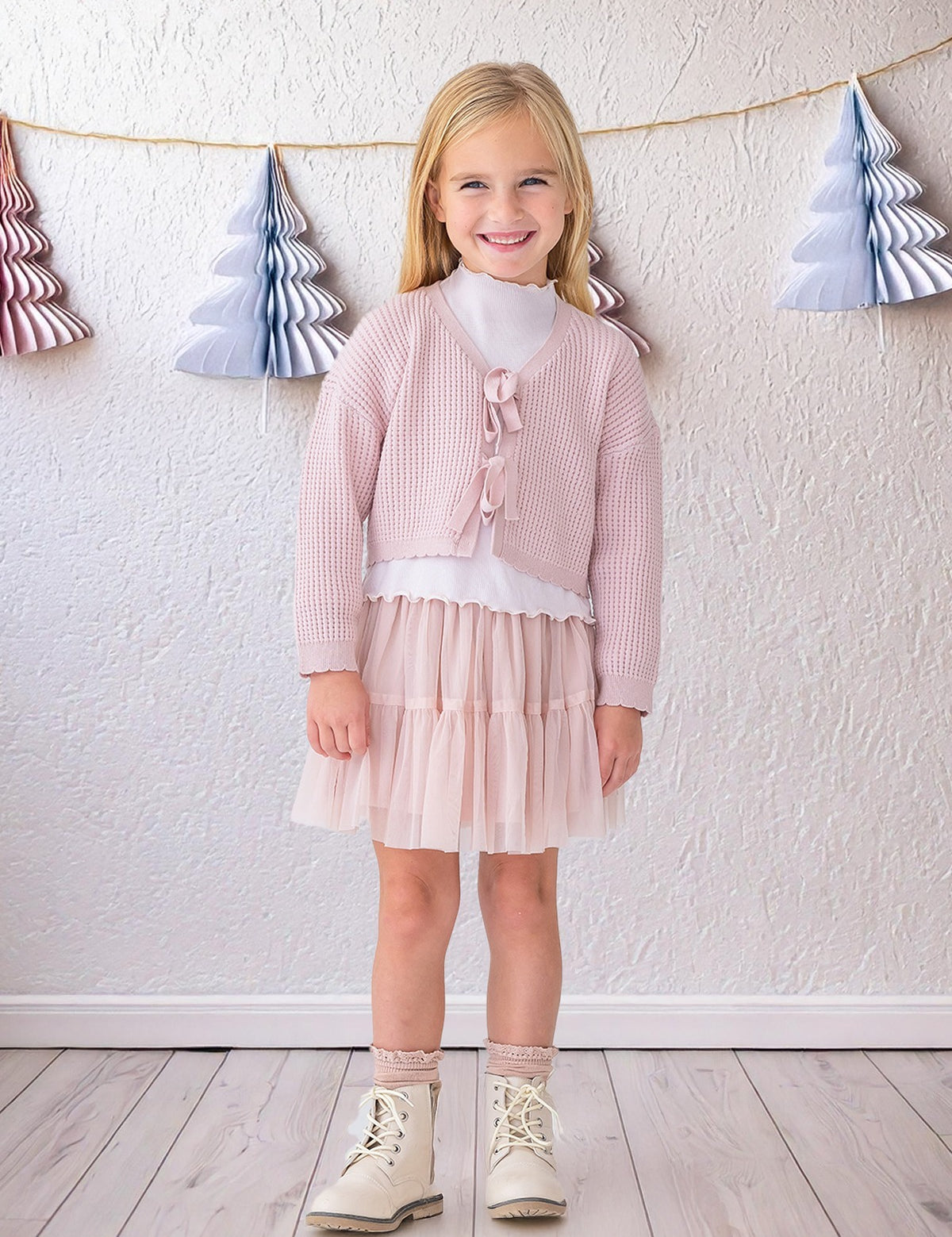 A young girl with blonde hair smiles indoors, wearing the Isobella and Chloe Ballerina Sweater over a white top, paired with a pale pink tulle skirt, sparkly socks, and white boots. A wall with paper Christmas tree garlands is behind her.