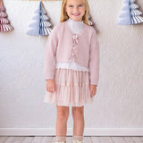 A young girl with blonde hair smiles indoors, wearing the Isobella and Chloe Ballerina Sweater over a white top, paired with a pale pink tulle skirt, sparkly socks, and white boots. A wall with paper Christmas tree garlands is behind her.