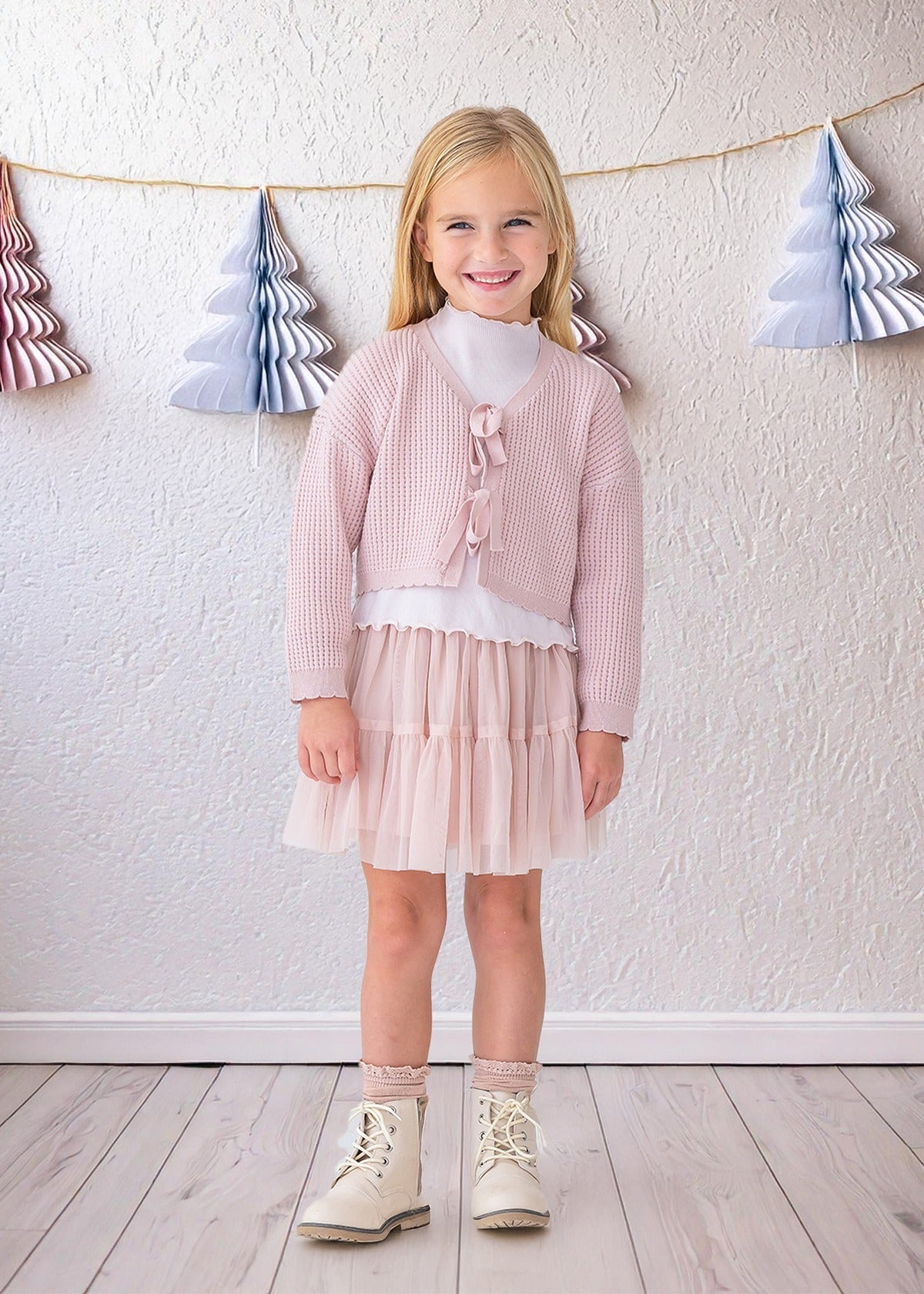 A young girl with blonde hair smiles indoors, wearing the Isobella and Chloe Ballerina Sweater over a white top, paired with a pale pink tulle skirt, sparkly socks, and white boots. A wall with paper Christmas tree garlands is behind her.